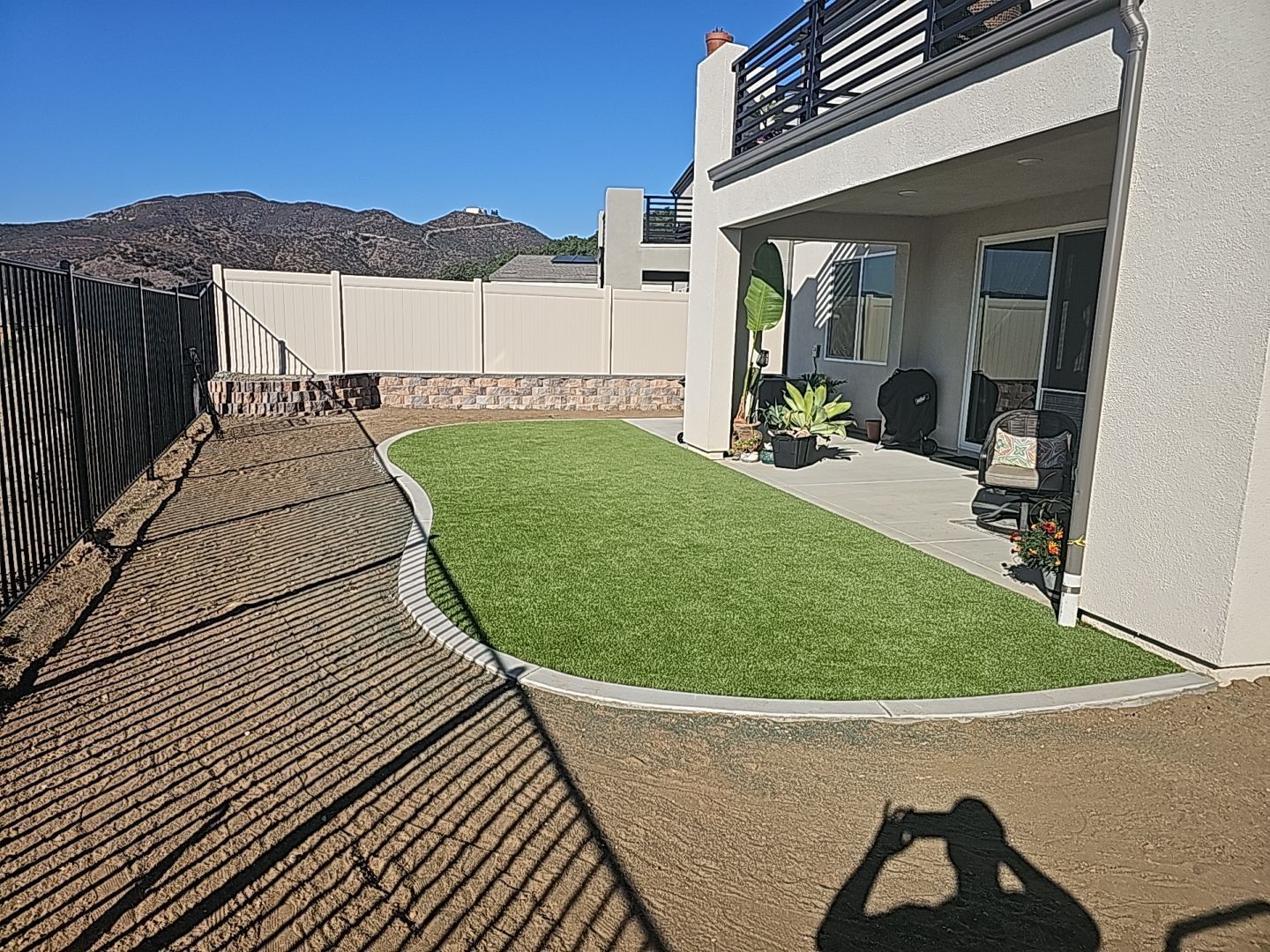 Backyard with green artificial grass, patio, fence, and bare dirt area. Shadow of a person taking a photo.