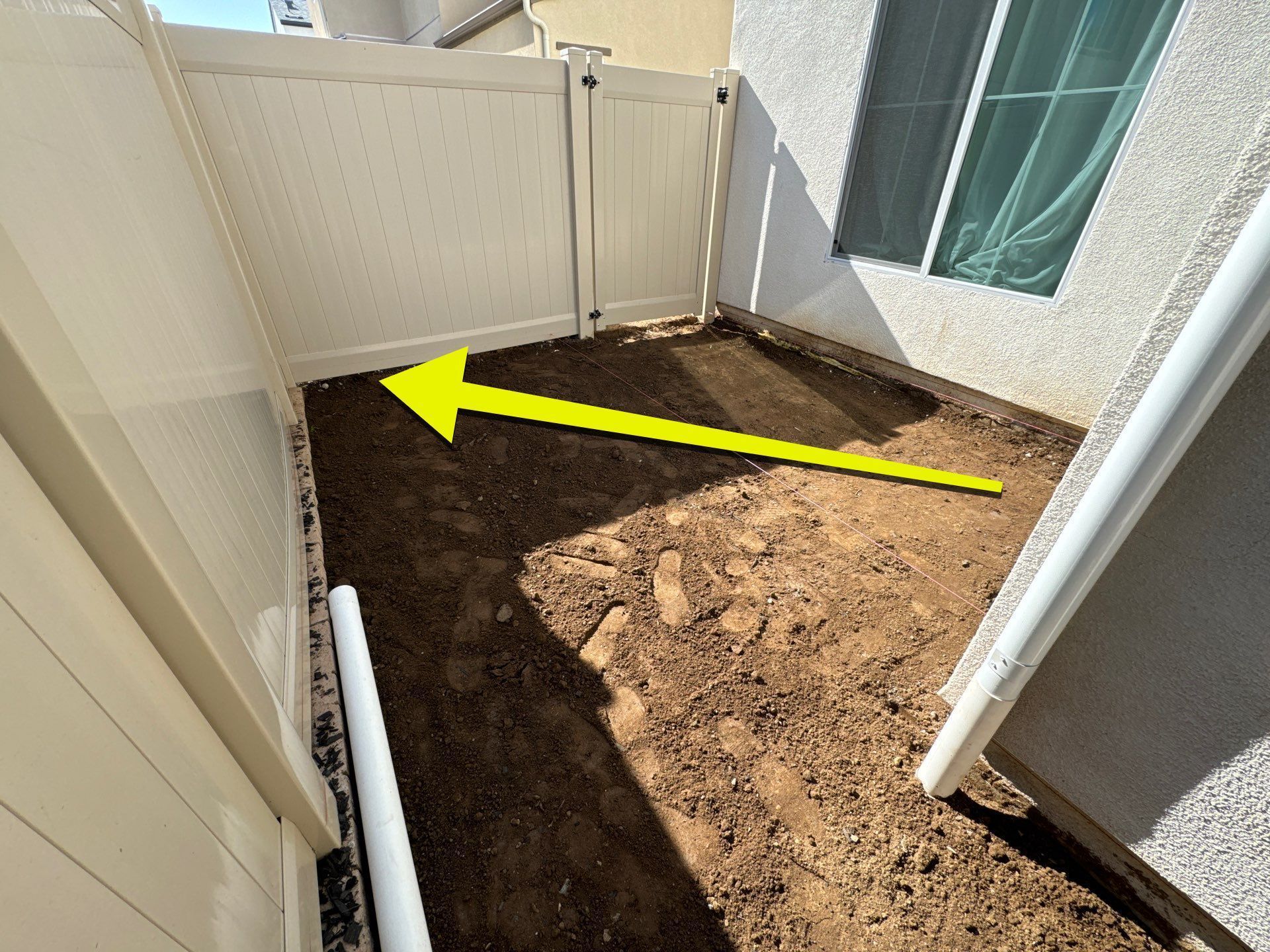 A narrow dirt path between white fences and a house. A yellow arrow points from left to right.