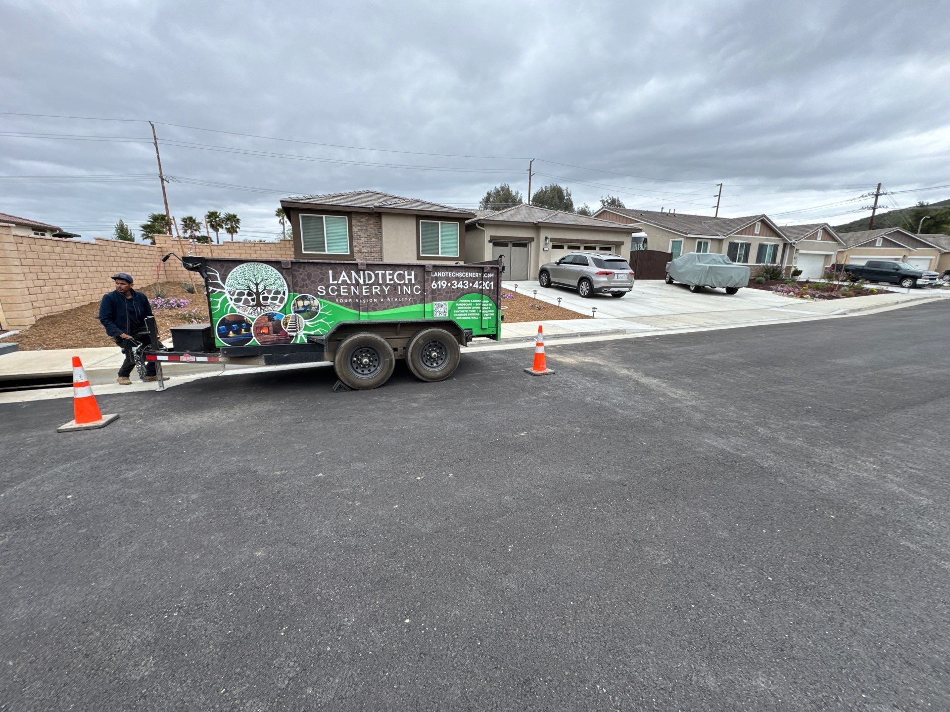 A man works near a trailer with landscaping company branding on a residential street. Cloudy sky.