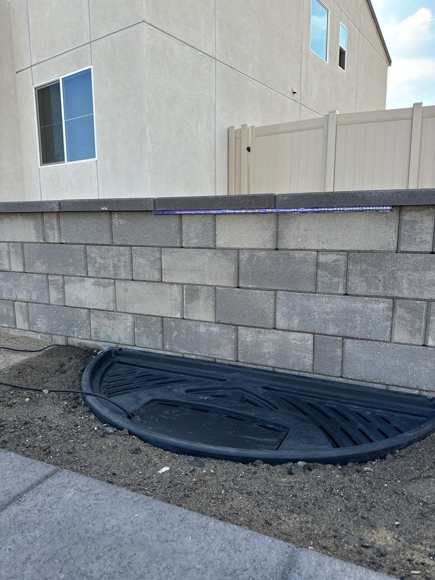 Black semi-circle well cover in front of a gray brick retaining wall, next to a building and paved sidewalk.