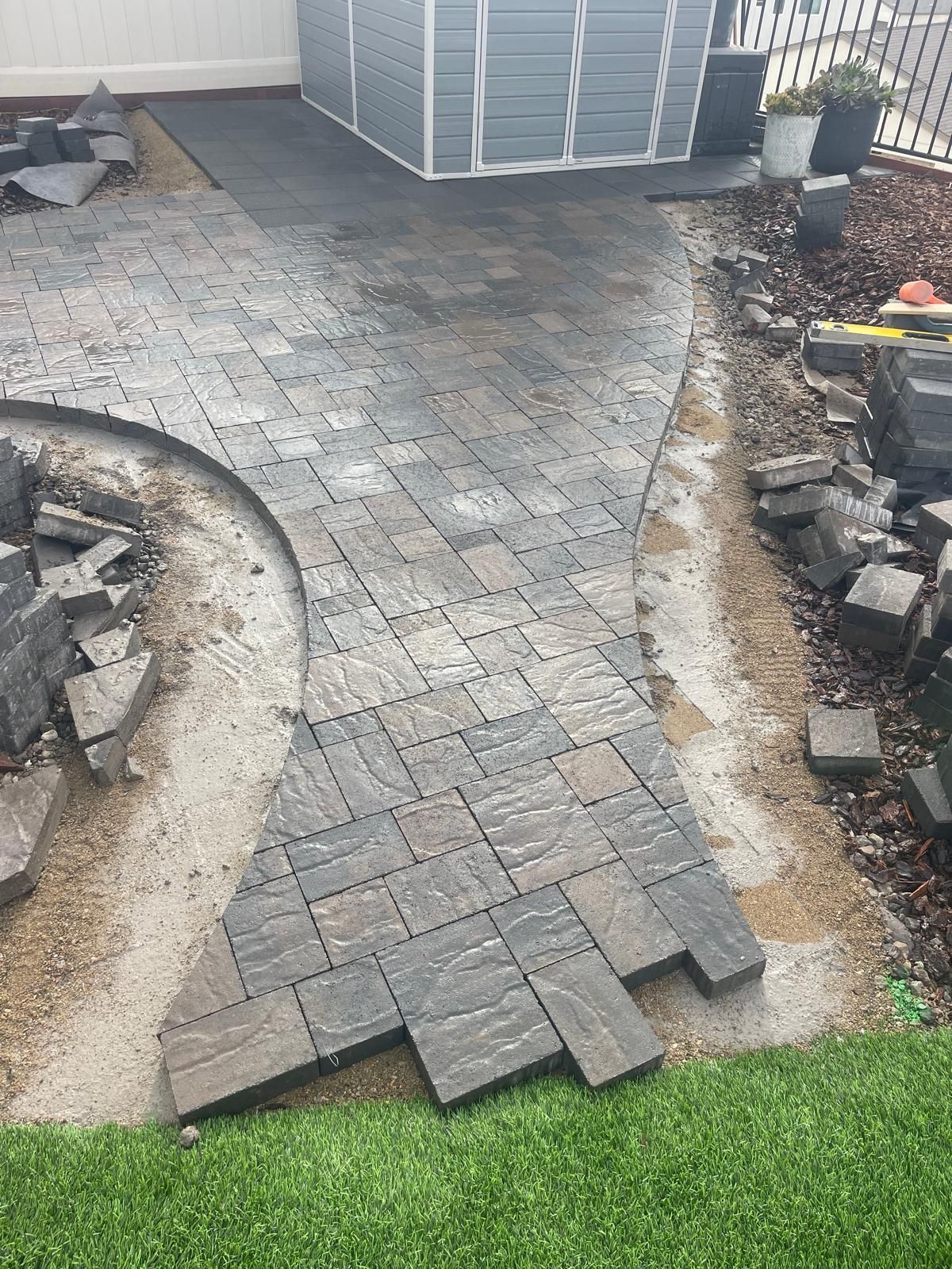 Pavers being laid in a garden, with a curved path and shed in the background.