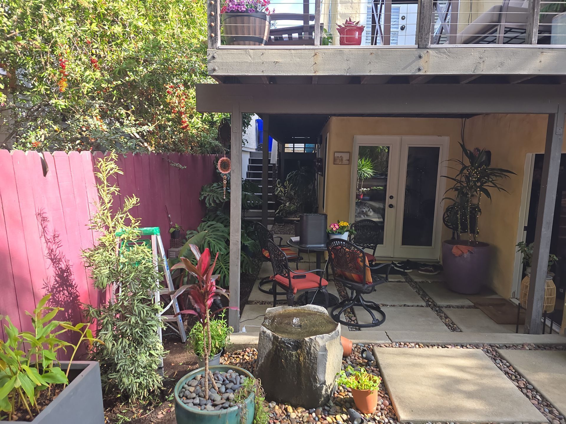 Backyard with patio, plants, and second-story deck. A fountain, table, and chairs sit on the stone patio.