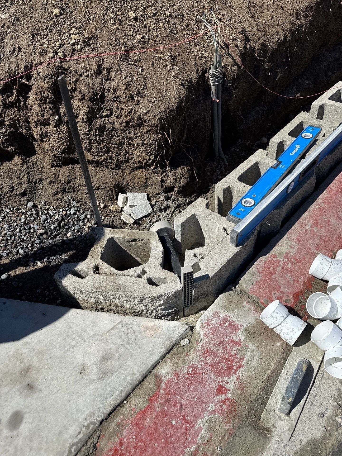 Construction site with cinder blocks, a level, and PVC pipes near a curb and dirt.