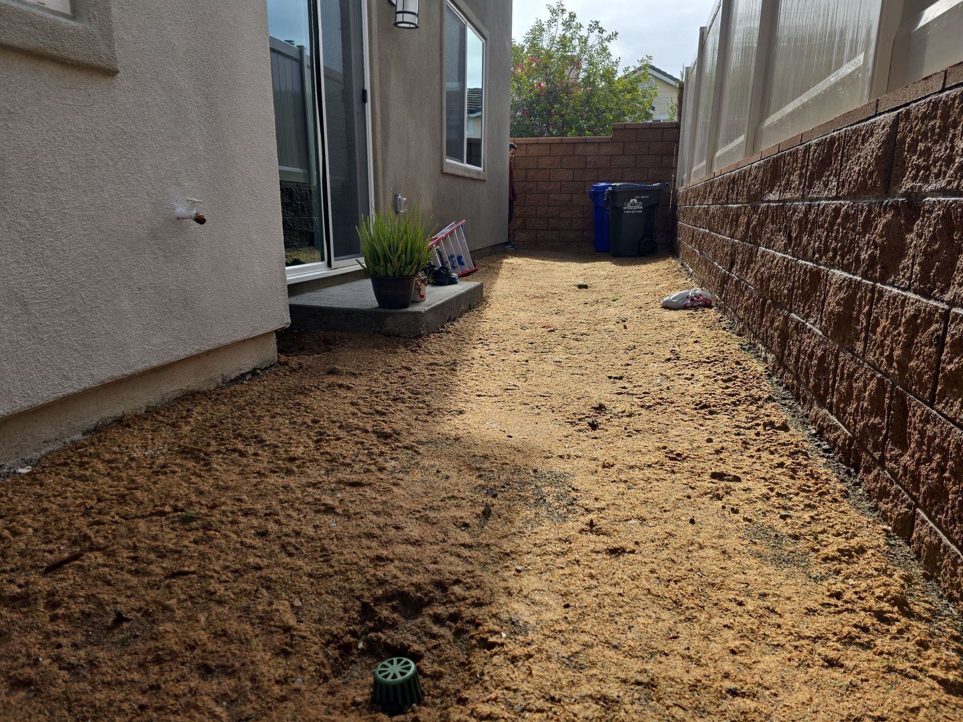 Backyard with dirt ground, brown retaining wall, and beige house wall.