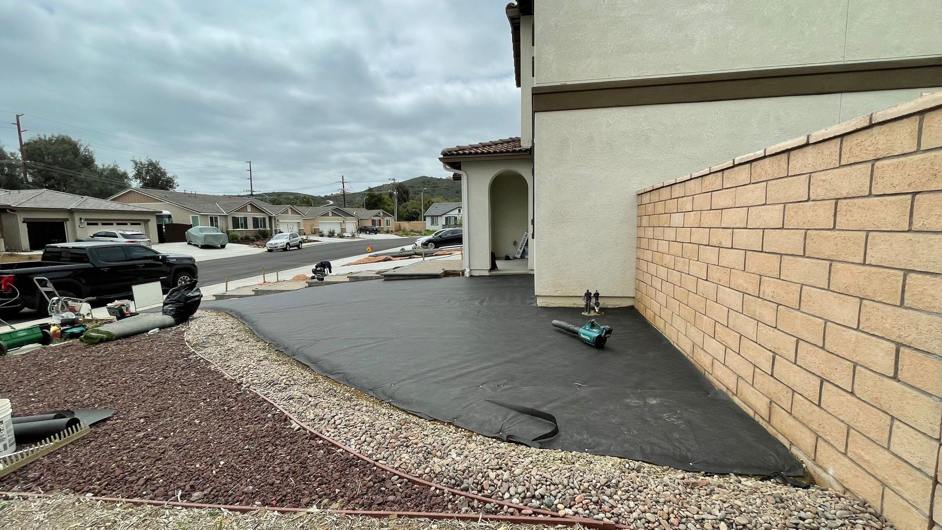 New concrete patio next to a beige-colored house and brick wall. Construction materials and a cloudy sky are visible.