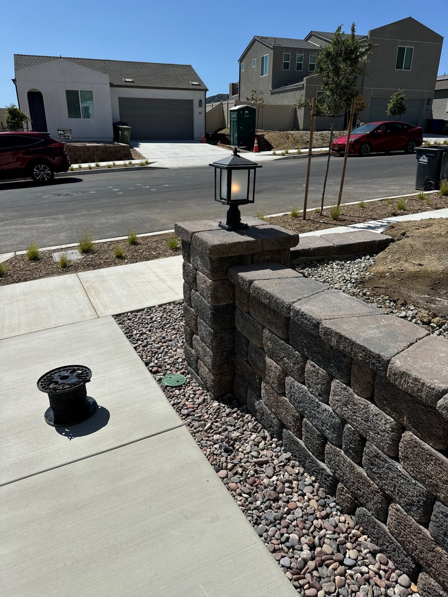 Stone retaining wall with a lamp post and small gravel bed. Houses and street in the background.