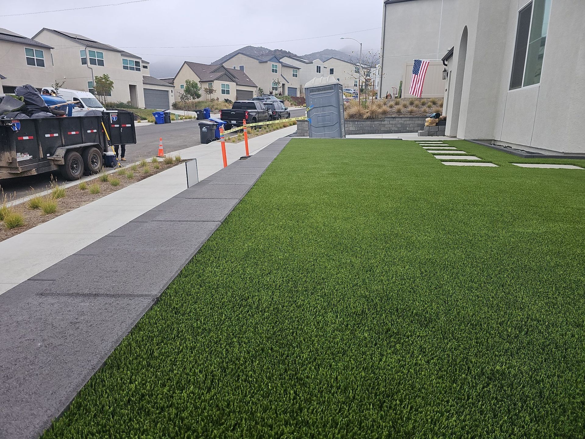 A green lawn borders a dark gray sidewalk and driveway in front of houses; a trailer and porta-potty are in the background.