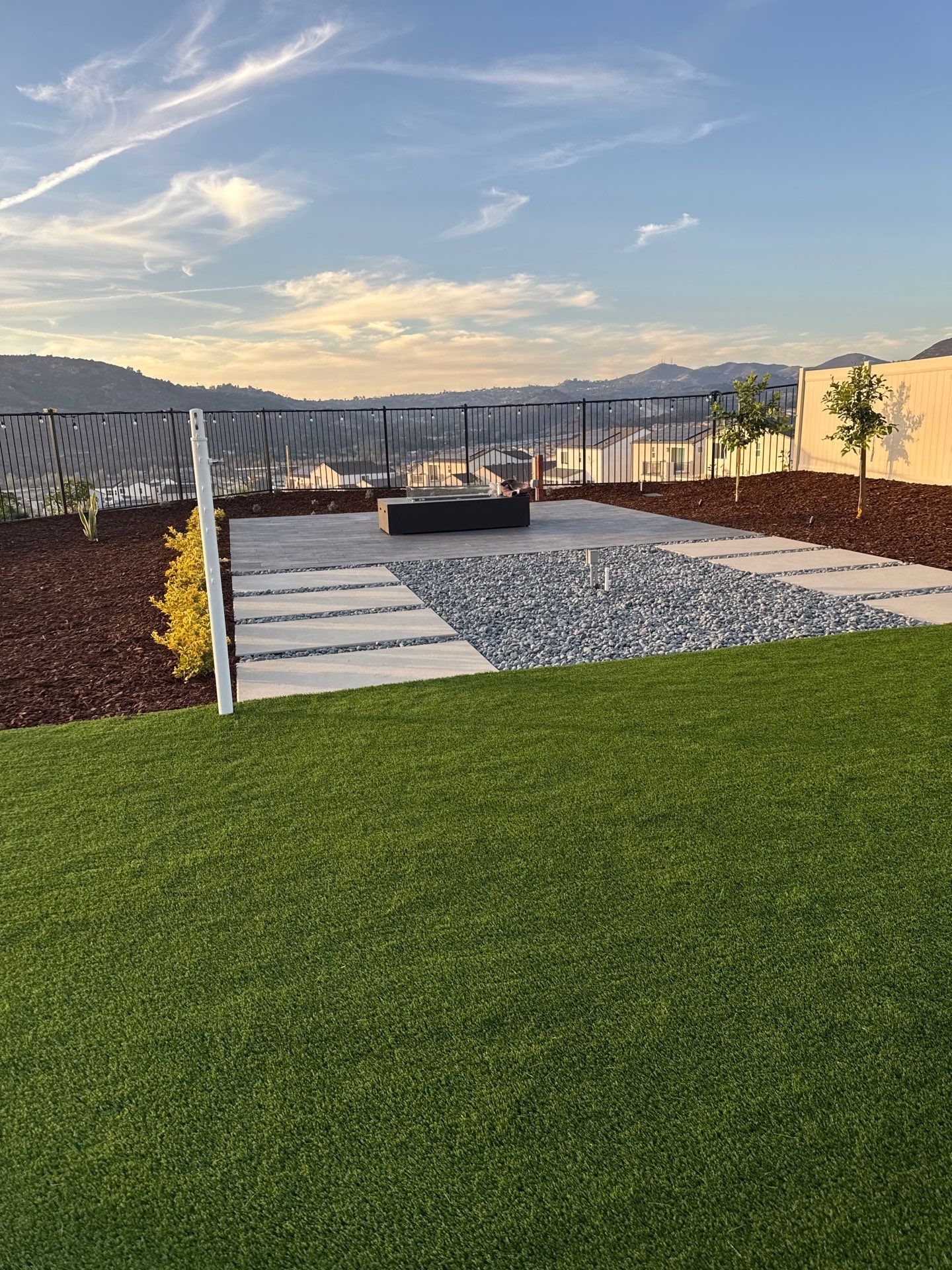 Backyard patio with fire pit, gravel, stepping stones, and green lawn; mountain view in background.