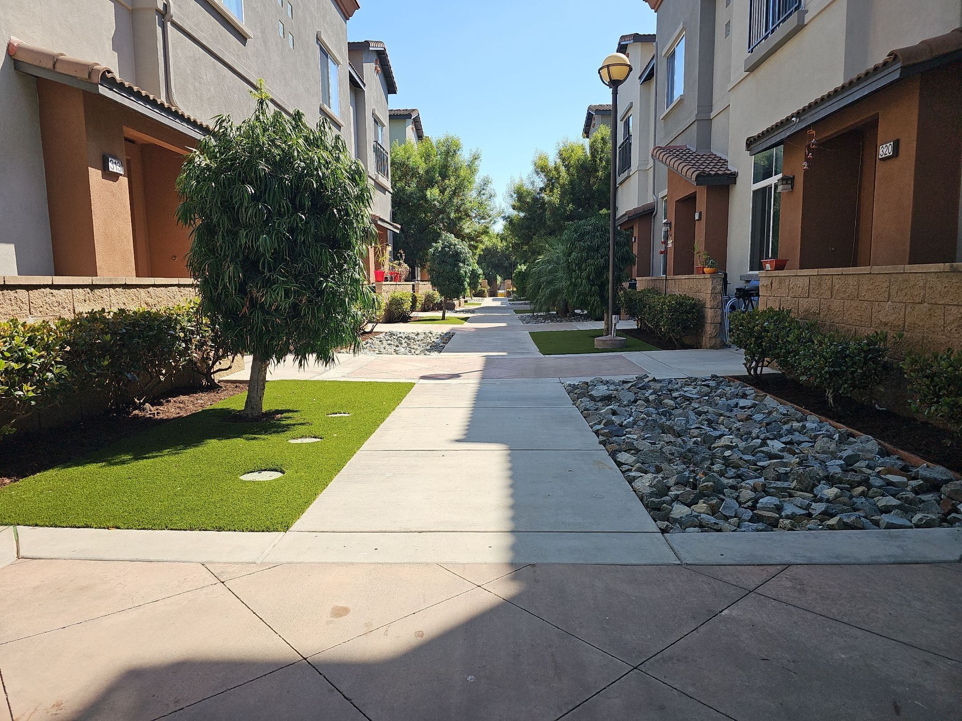 Sidewalk in a residential area. Beige buildings with covered entrances line the sides of the path. Landscaping includes grass, trees, and rocks.