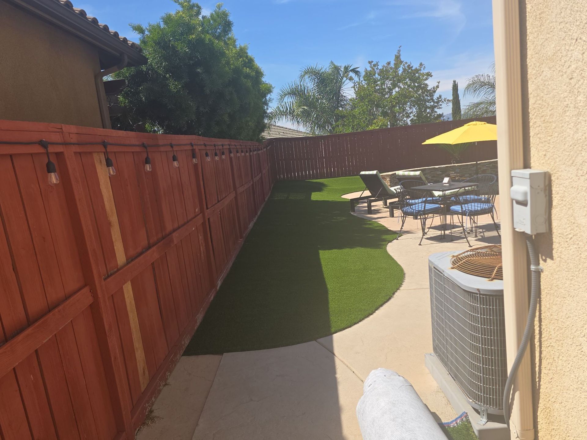 Backyard with red fence, green lawn, patio furniture, and string lights. Sunny day.