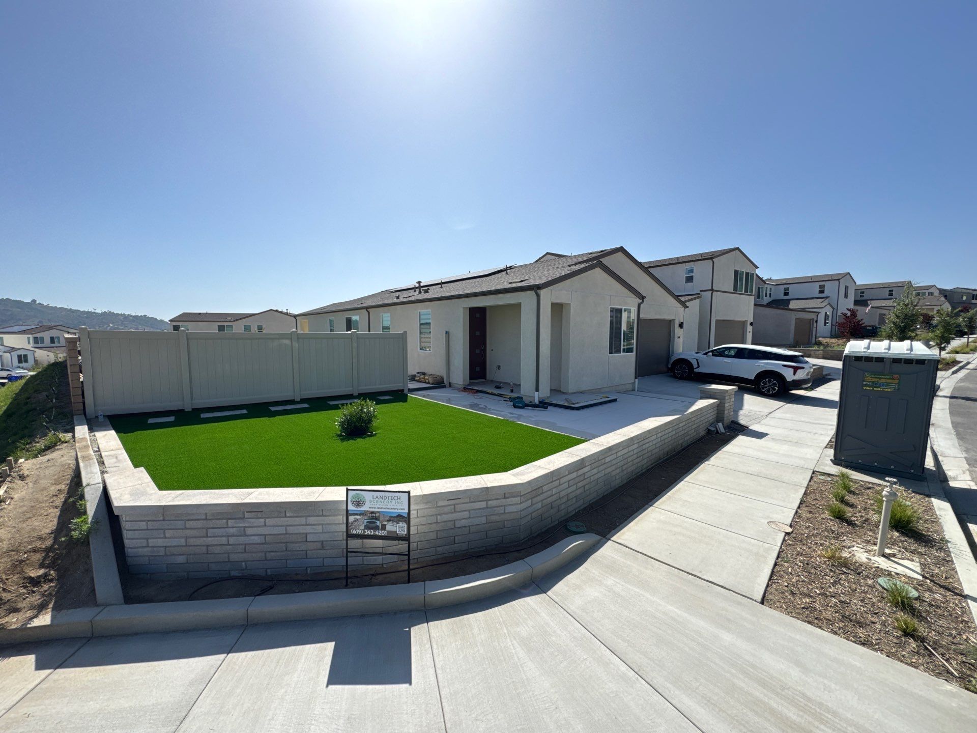 A light beige house with a concrete driveway, retaining wall, and green lawn. Clear blue sky.