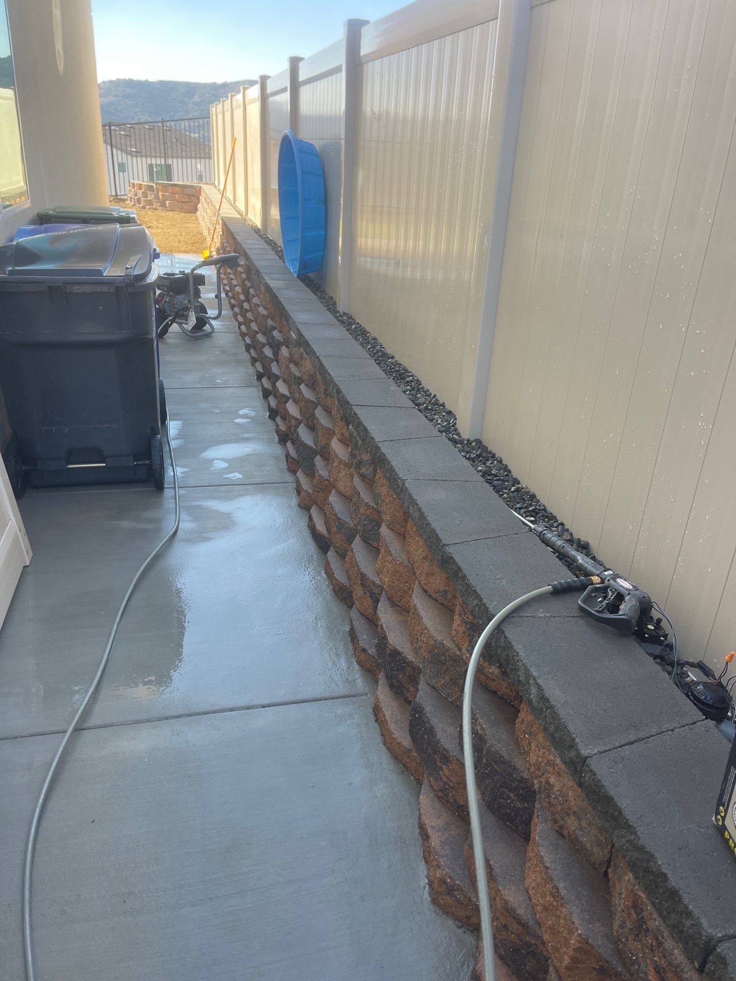 A wet, gray patio with a brick retaining wall along a white fence. A trash can and hose are visible.