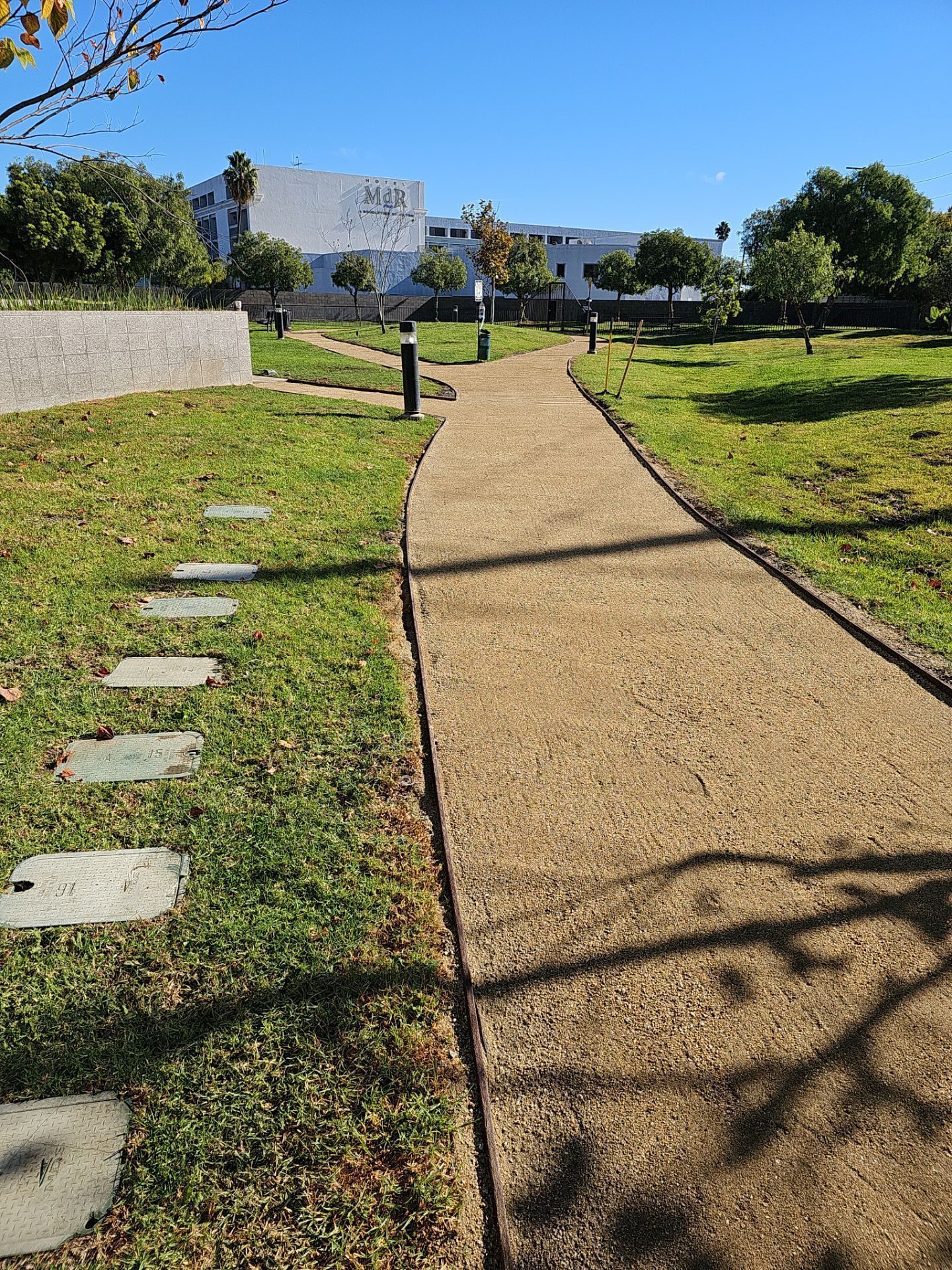 Gravel path winds through a park with grass, trees, and stone steps, towards a white building under a blue sky.