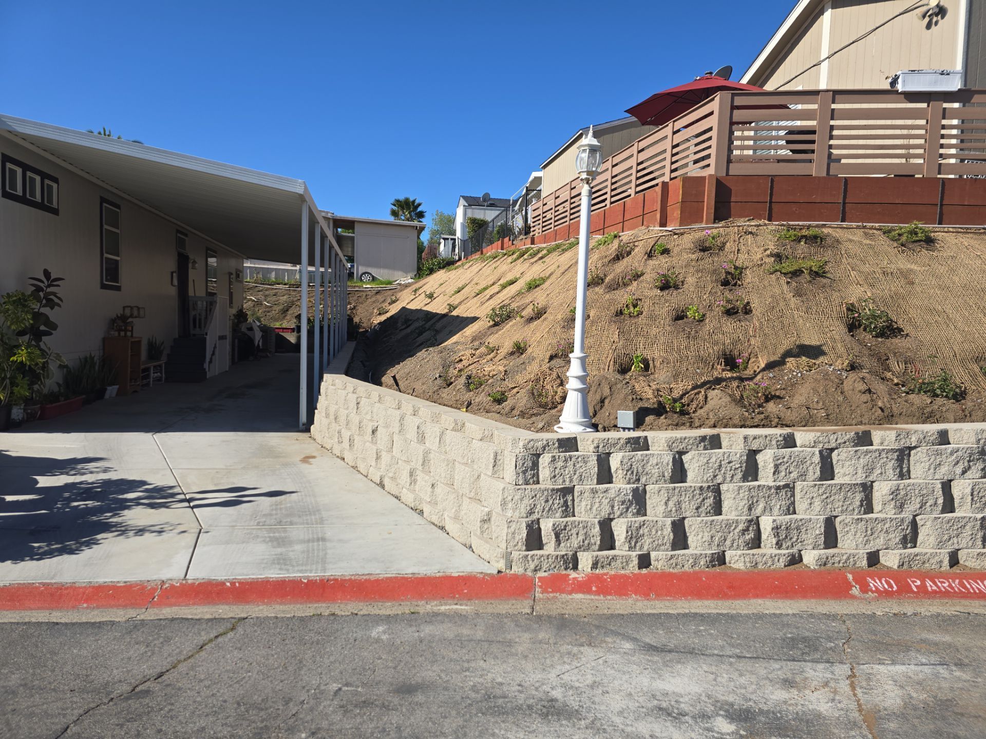 Street view: retaining wall, sloping hillside, houses, a carport, and a blue sky.