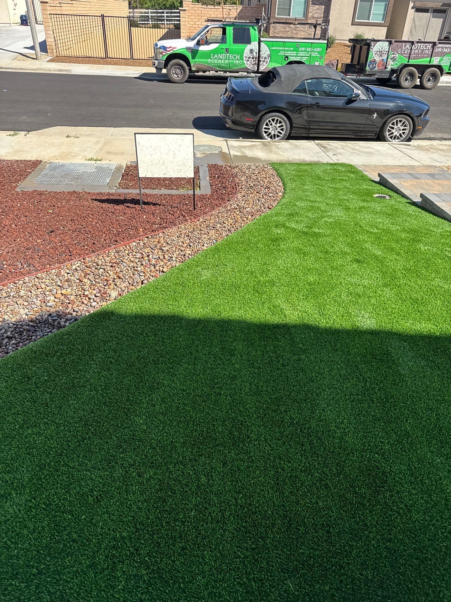 Green artificial turf with red mulch border and a black car parked on a street.