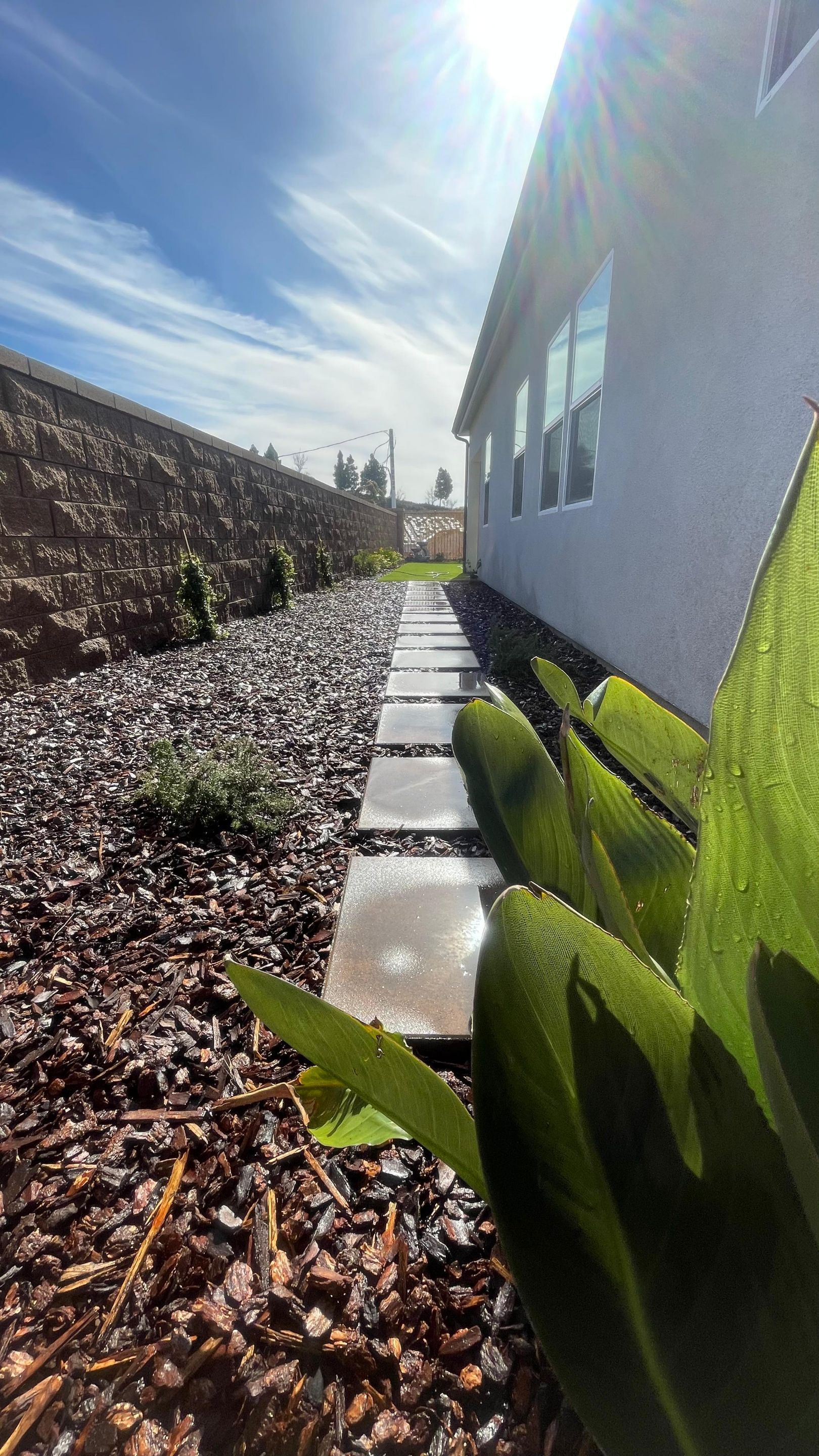 Stone path alongside a house, with a garden bed of dark rocks and plants under a bright sky.
