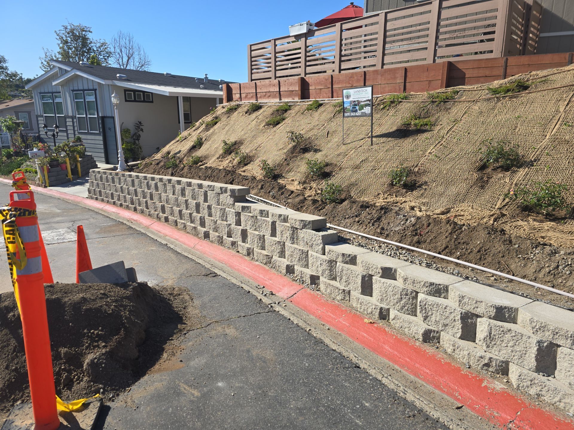 Retaining wall made of gray blocks along a road. Dirt hill with young plants behind it.
