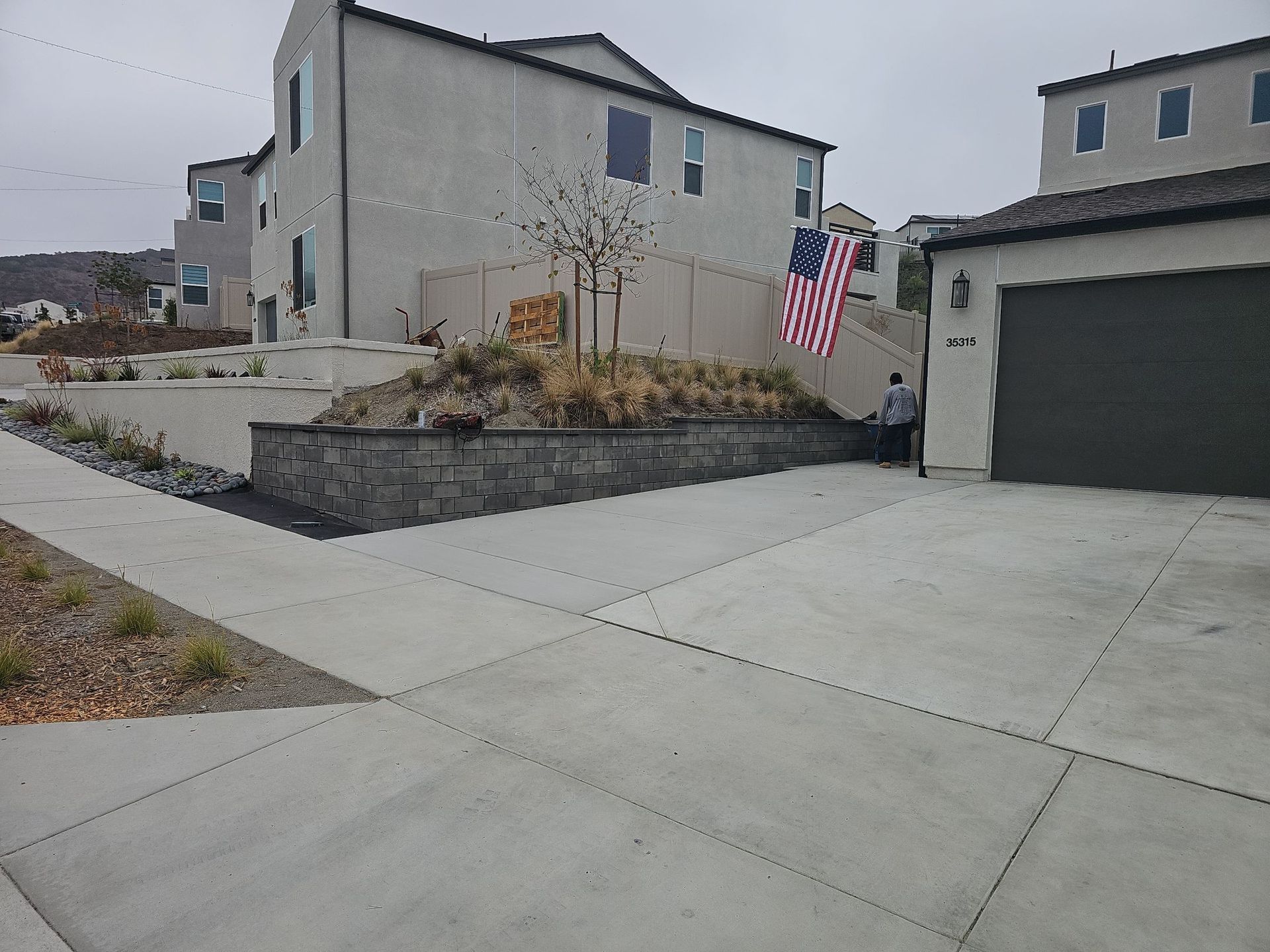 Residential street scene: houses, driveway, retaining wall with plants, American flag. Overcast sky.