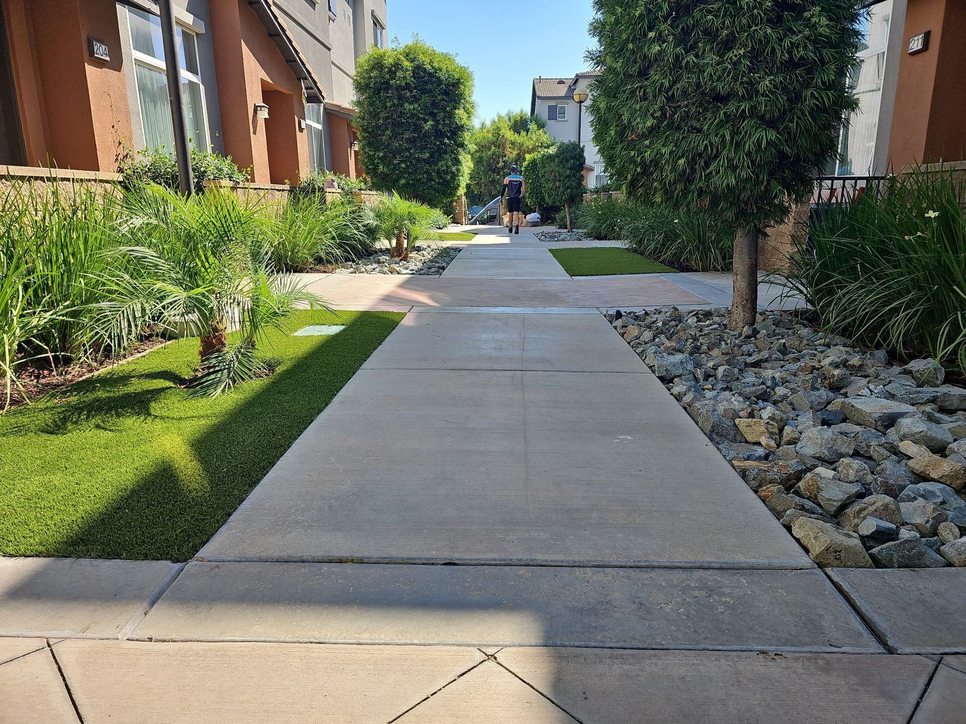 A paved walkway with a person walking towards residential buildings, flanked by greenery and rock landscaping.