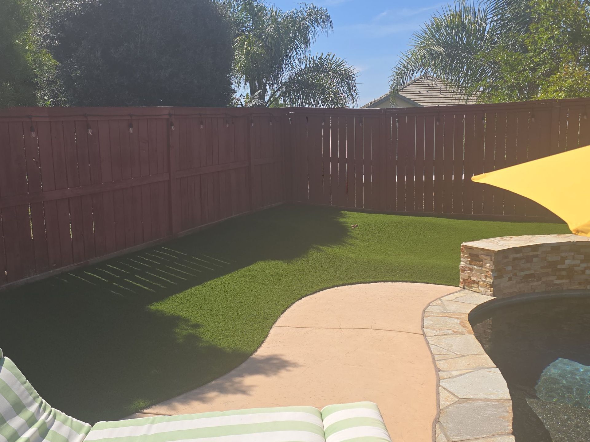 Backyard with green lawn, brown fence, and a curved stone pathway leading to a pool with a yellow shade.