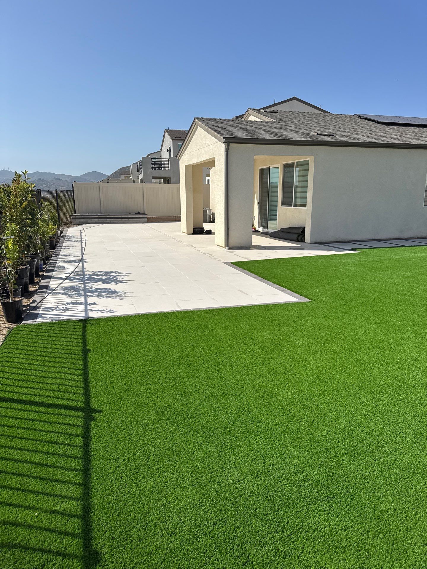 Backyard with green turf, concrete patio, beige house, and potted trees under a blue sky.