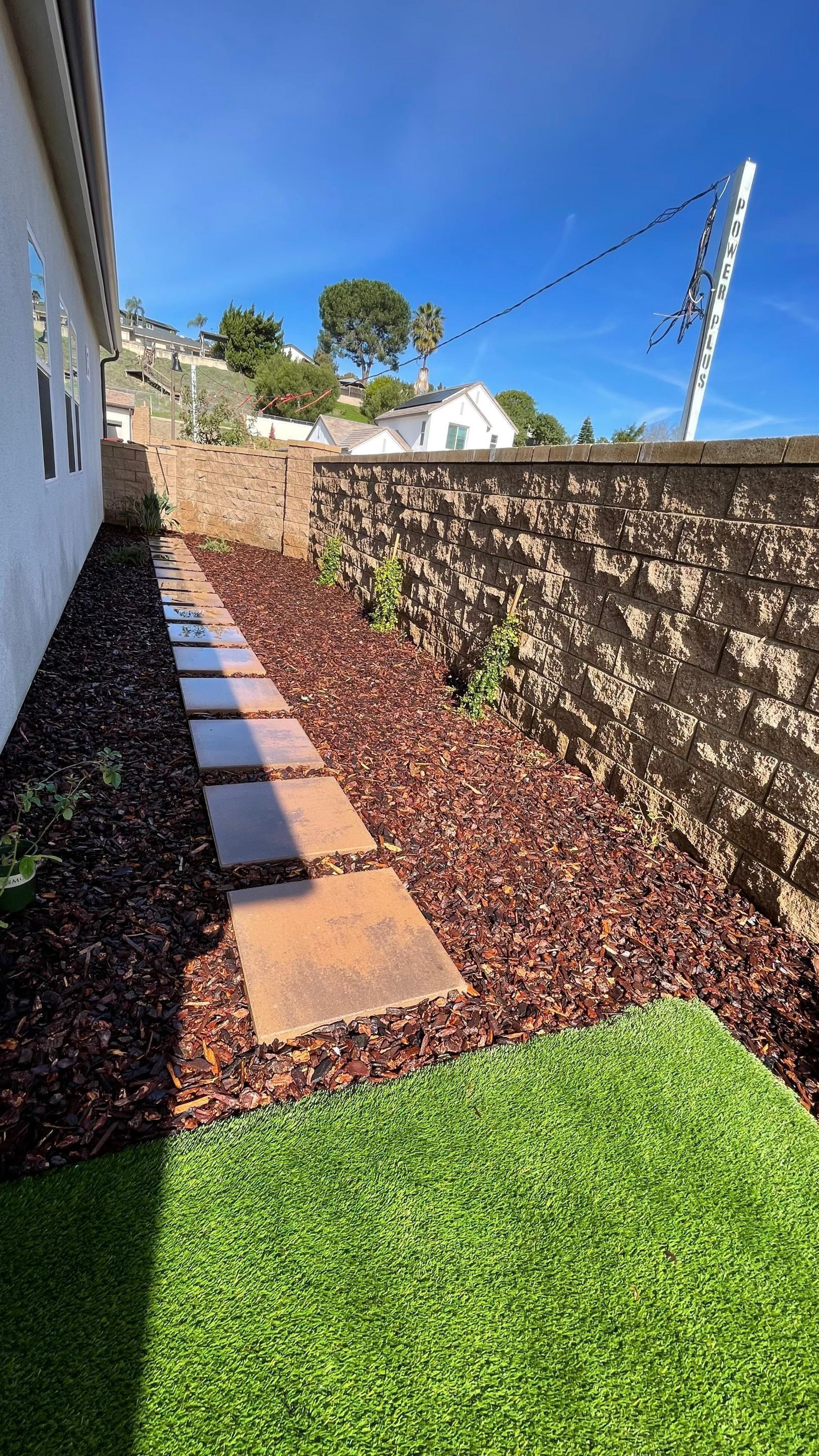 Stone path leads through a garden bed with mulch and artificial turf alongside a stone wall and white building under a blue sky.