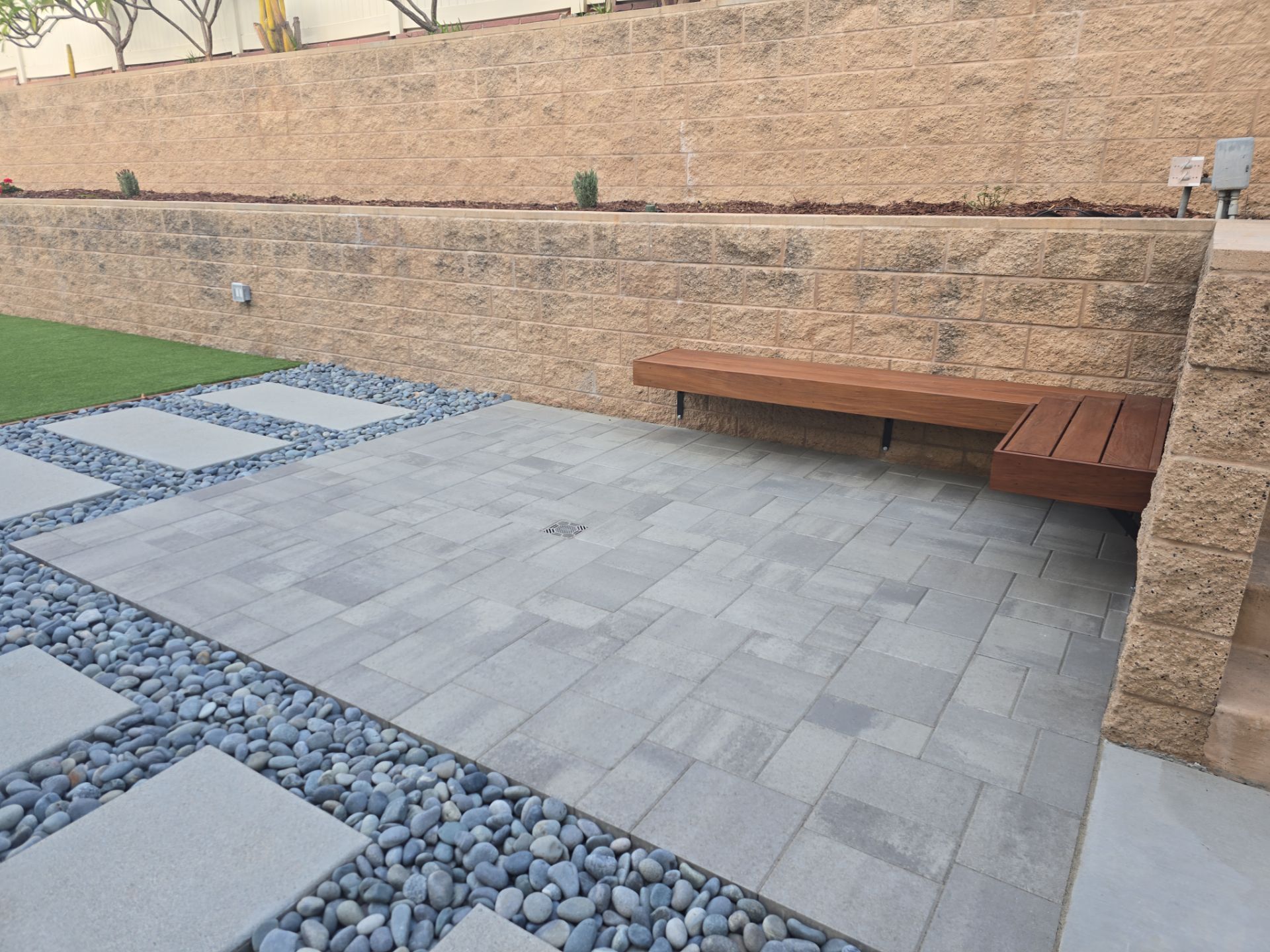 Patio with gray pavers, black pebbles, and a wooden bench against a retaining wall.