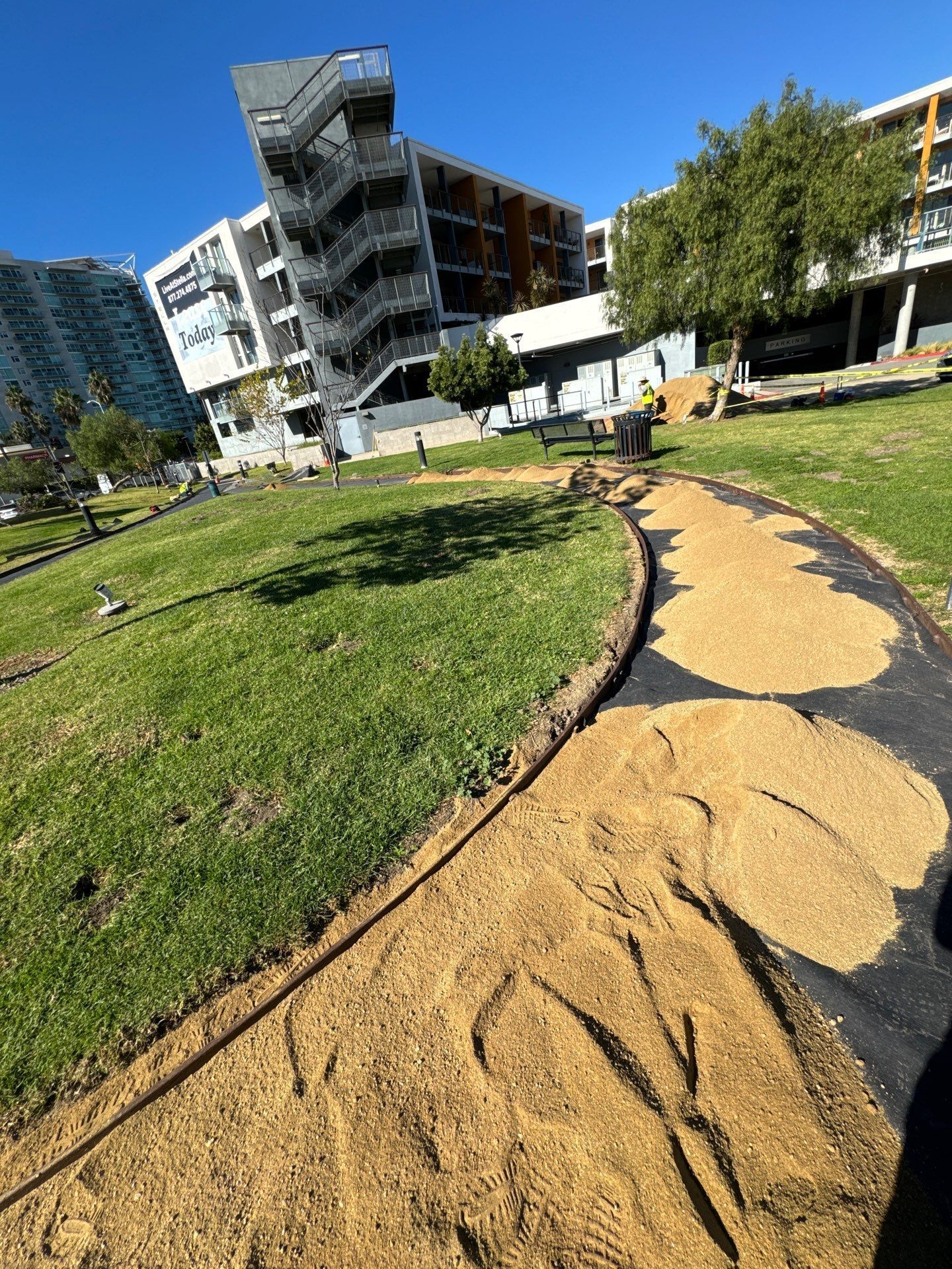 A walking path with tan mulch is being prepared in a green grassy area near apartment buildings.
