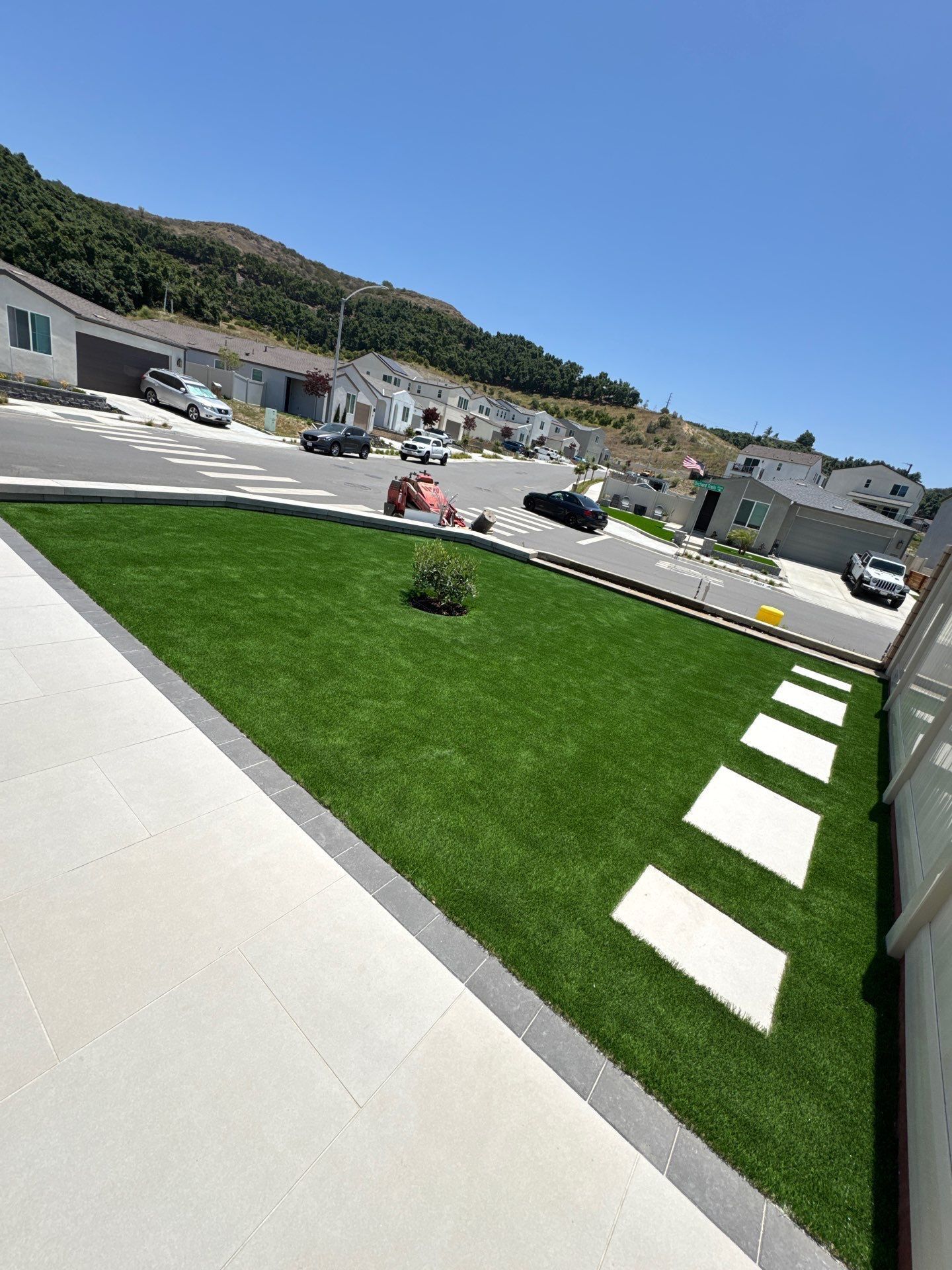 Green lawn with stone pathway next to a concrete patio, street, and houses on a sunny day.