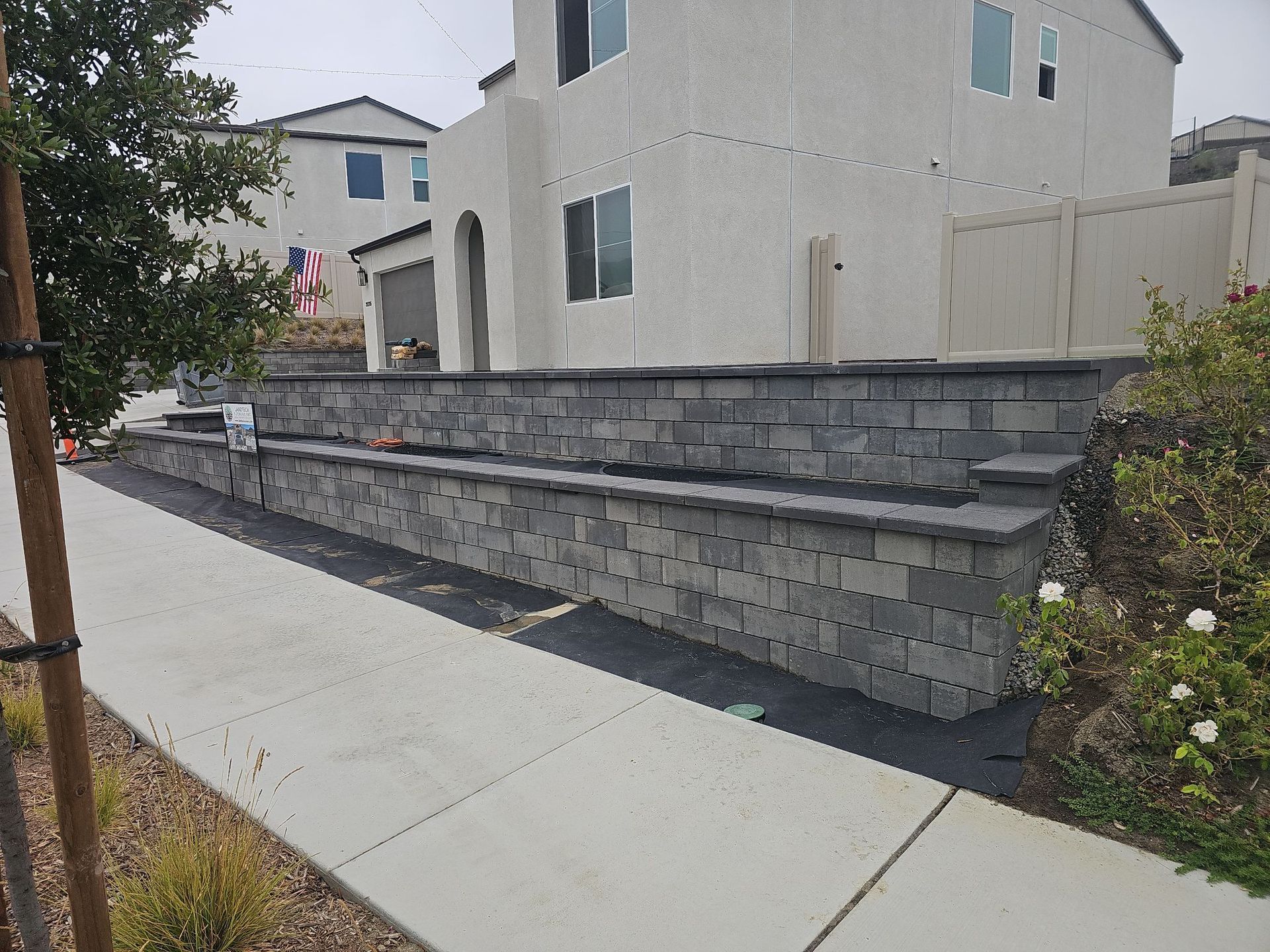 Gray stone retaining wall in front of a light-colored two-story house, with a sidewalk in foreground.