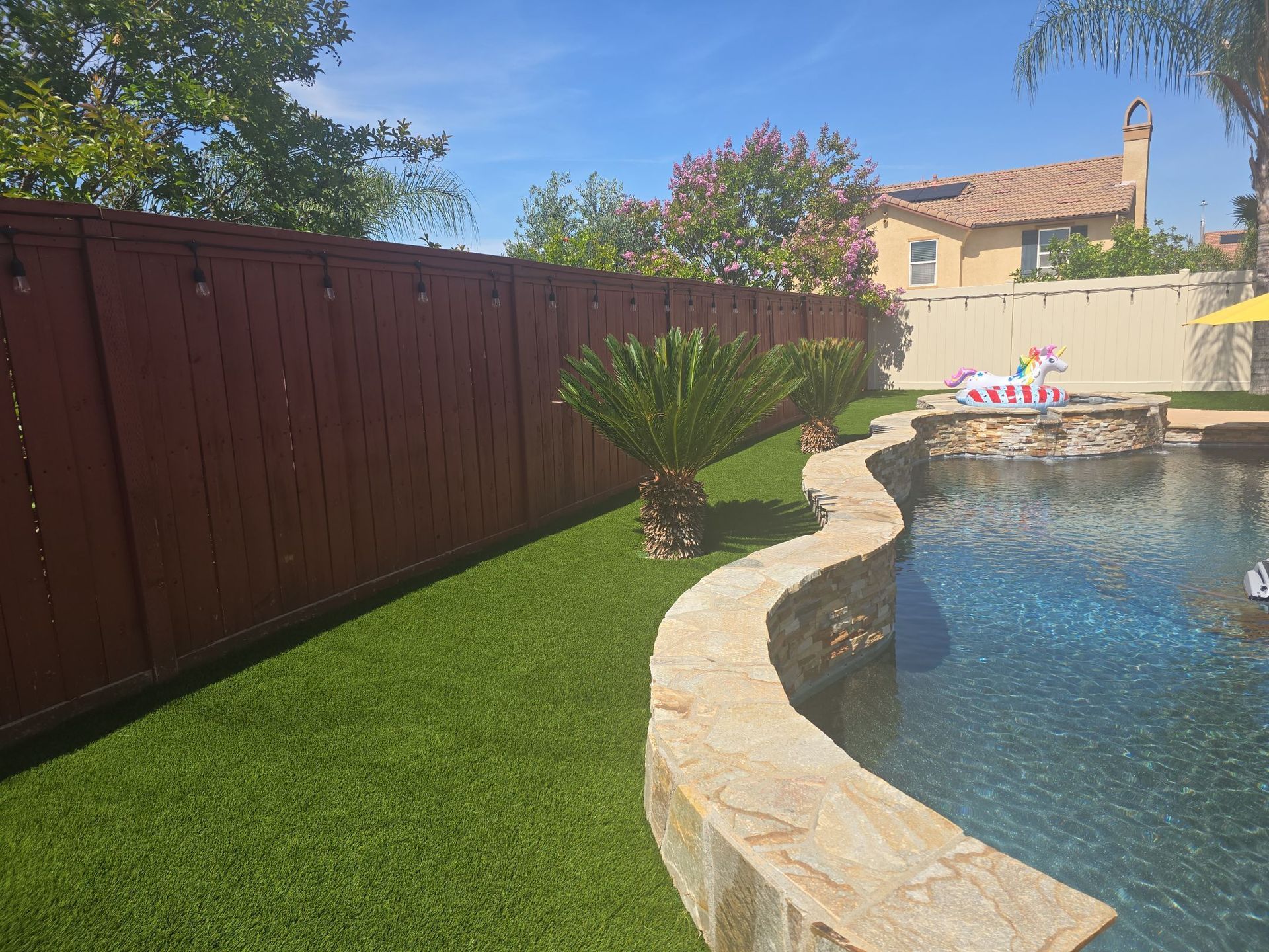 A backyard with a pool, brown fence, and green artificial turf under a blue sky.
