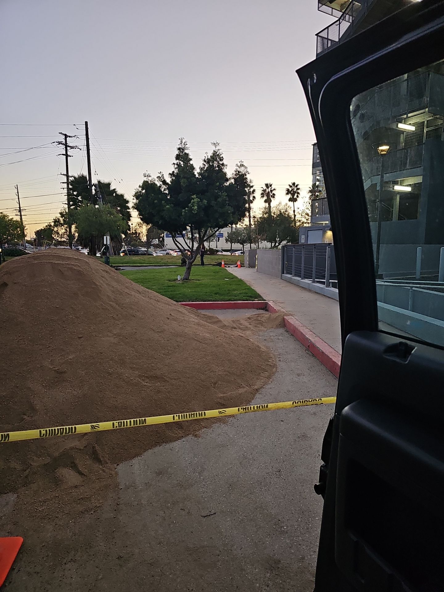 A pile of dirt next to a sidewalk with caution tape and an open car door. Evening setting.