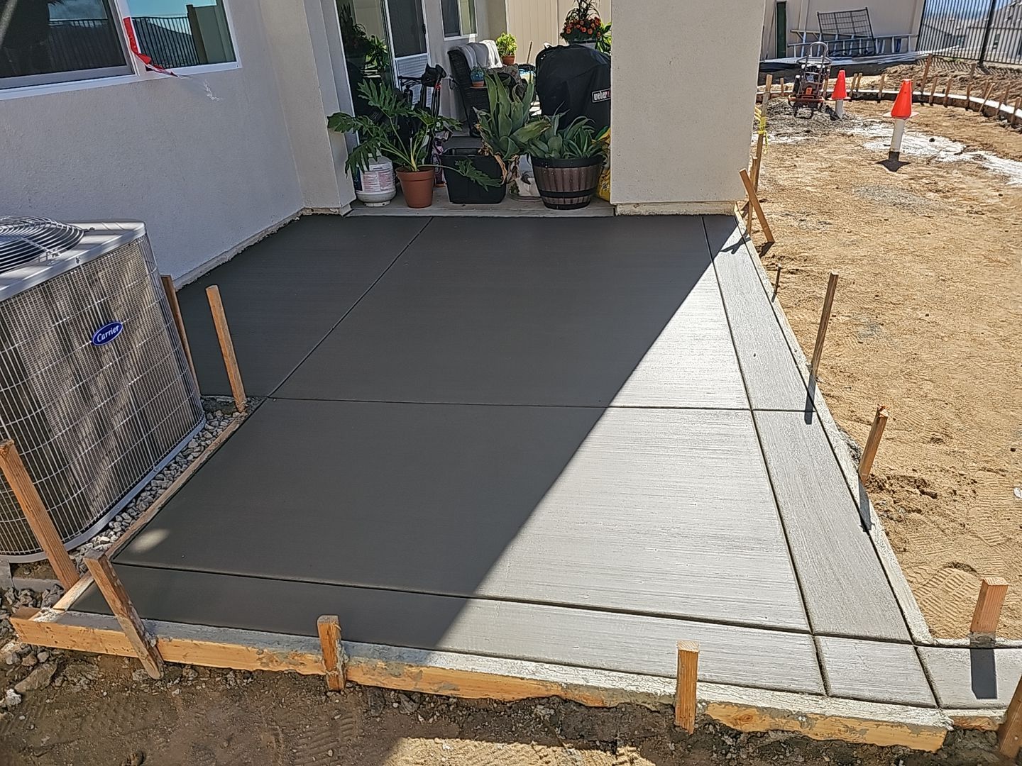 Newly poured concrete patio next to a house with plants and framing lumber visible.