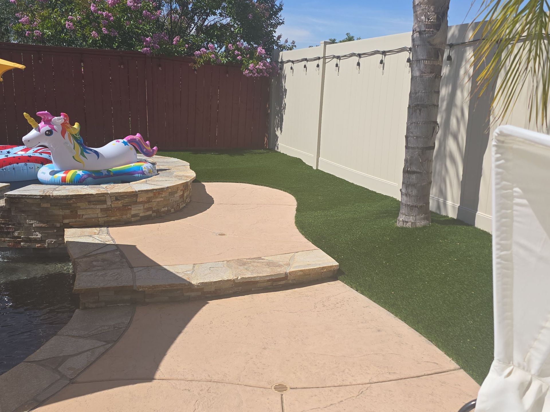 Patio with steps, artificial grass, a pool, and decorative fence. A unicorn pool float is present.