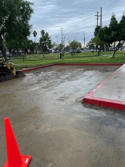 Construction site at park: freshly poured concrete area with red curb, orange cone, and small excavator. Cloudy sky.
