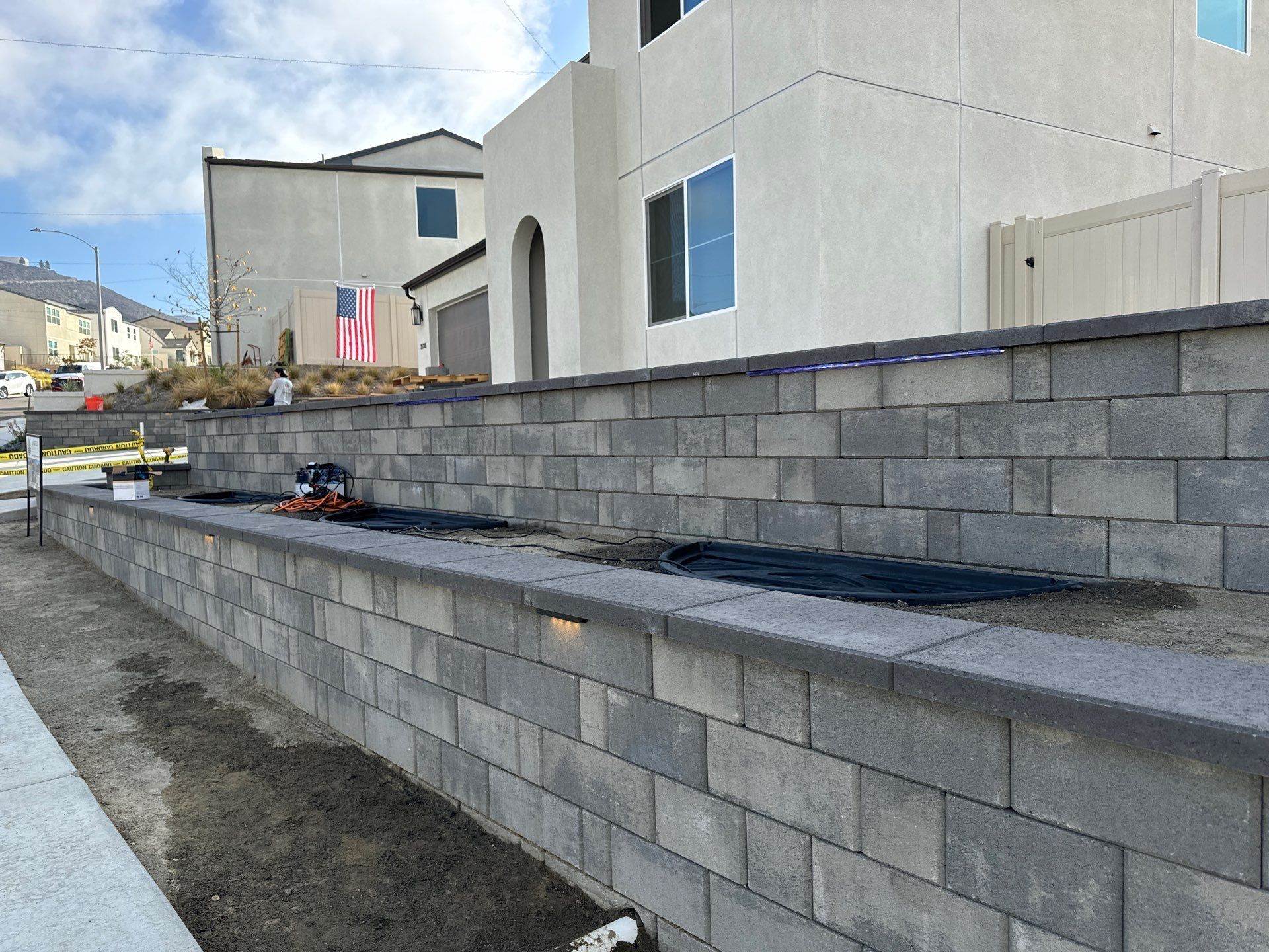 Gray block retaining wall along a sidewalk, with a light-colored house in the background.