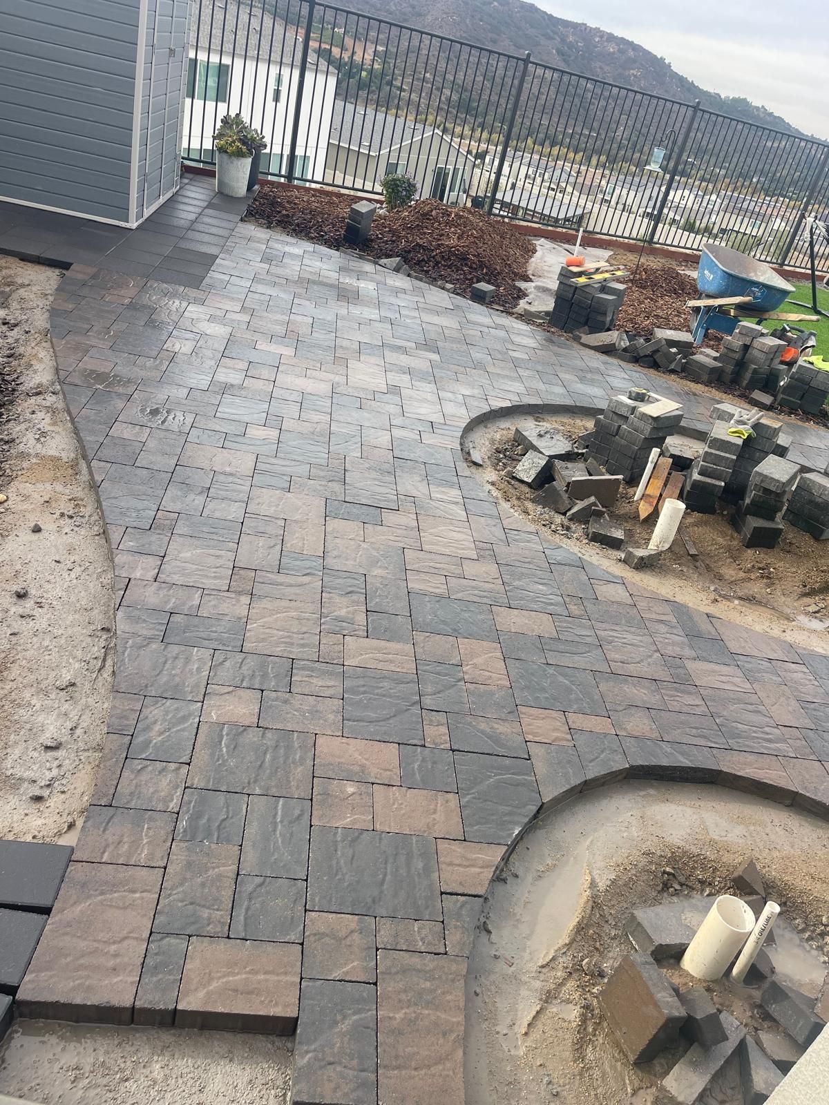Brick patio under construction, with dark and brown pavers. White pipes and landscaping materials are nearby.