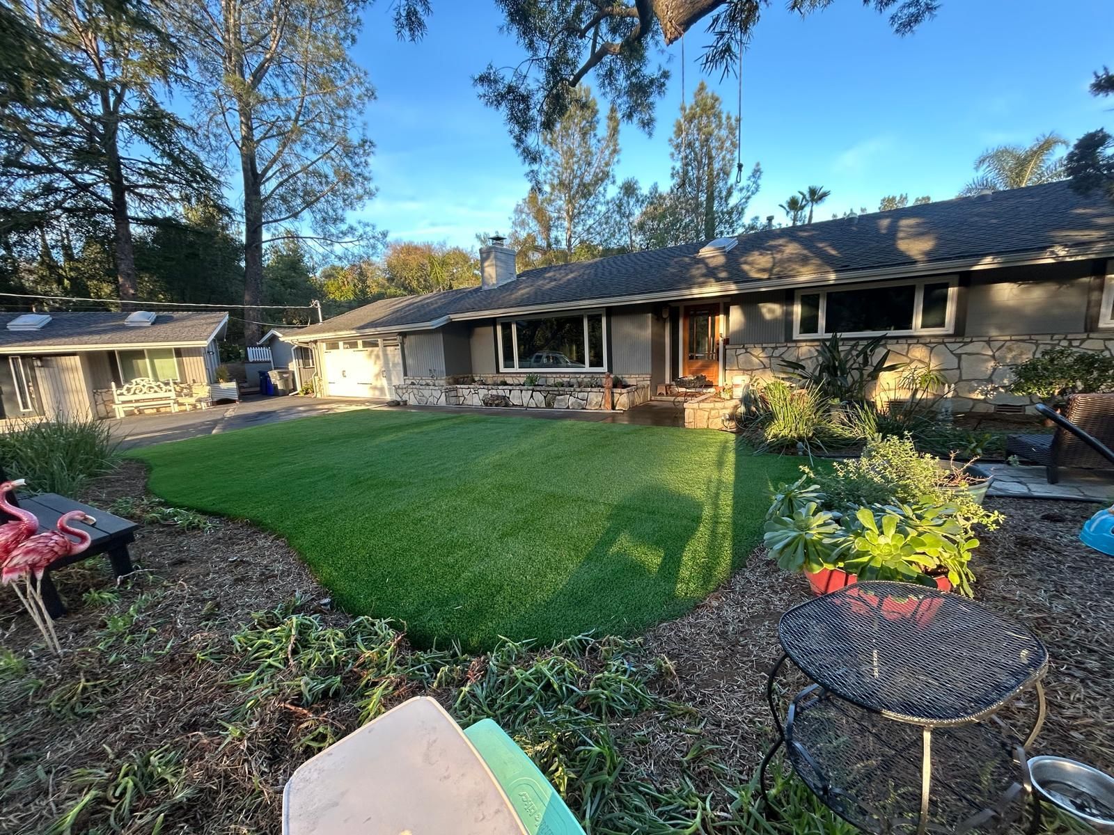 Single-story home with lush green lawn, mature trees, and landscaping under a blue sky.
