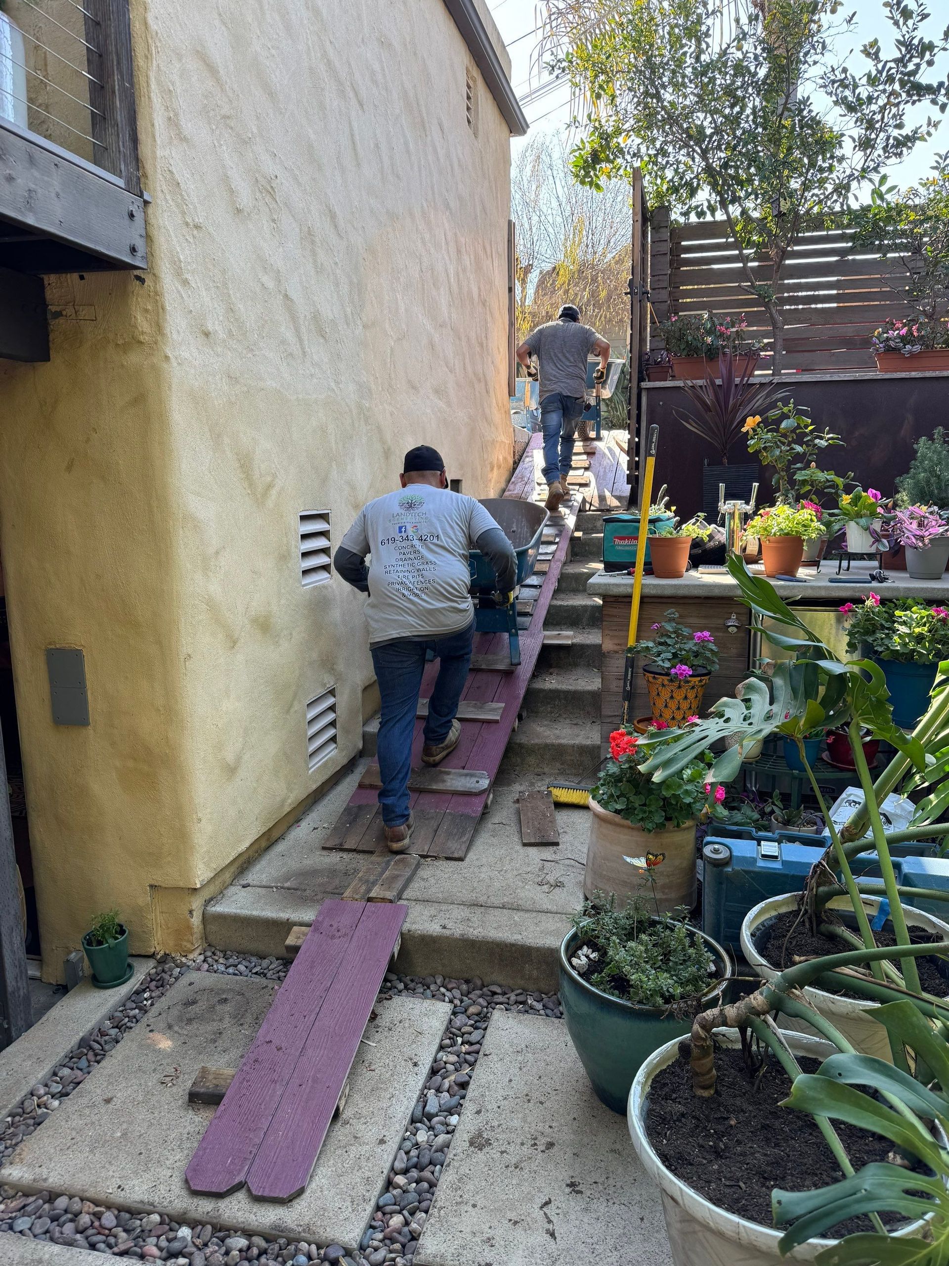 Two people carrying an object up a ramp built over steps alongside a stucco building, with potted plants.