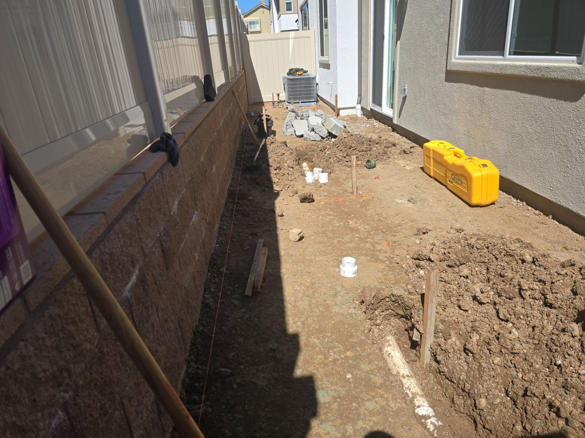 Backyard trench with pipes, dirt, and retaining wall. A yellow container sits nearby.