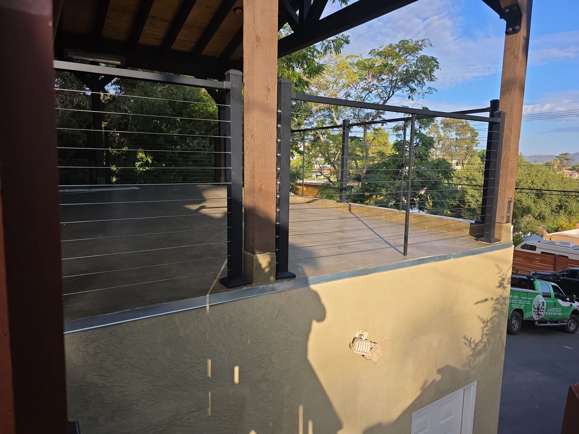 Outdoor deck with brown wooden posts, black metal railing, and neutral-colored stucco wall. Trees in the background.