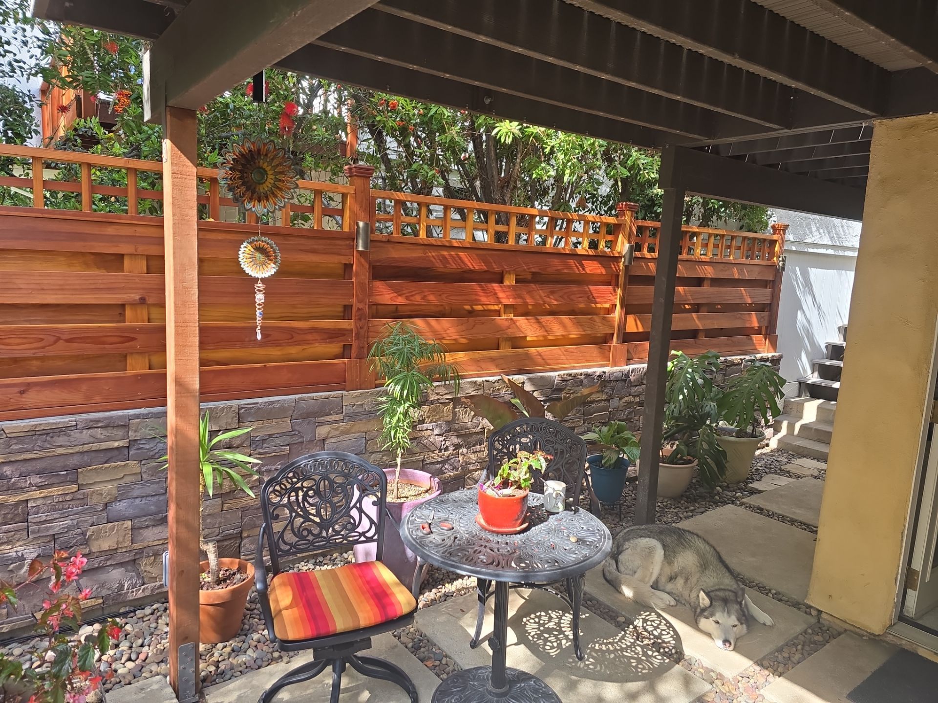 A small outdoor patio with a metal table and chair, with a wooden fence and plants.