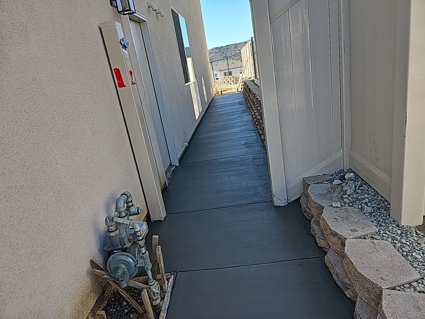 Narrow concrete walkway between a stucco wall and a white fence, leading to a backyard.
