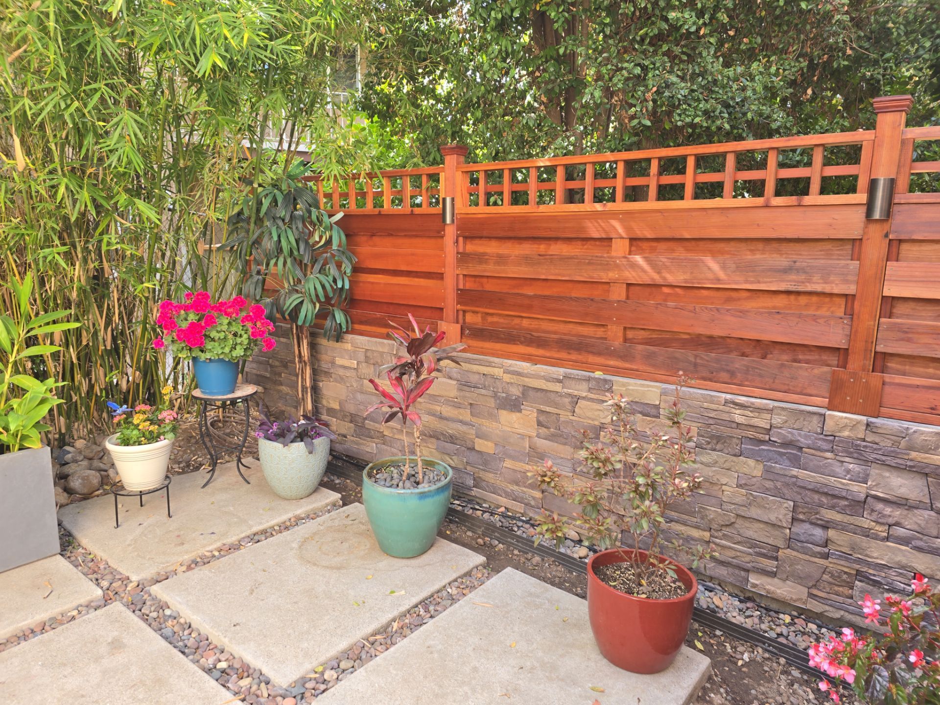 Patio with potted plants on stone pavers, next to a bamboo and wood fence.