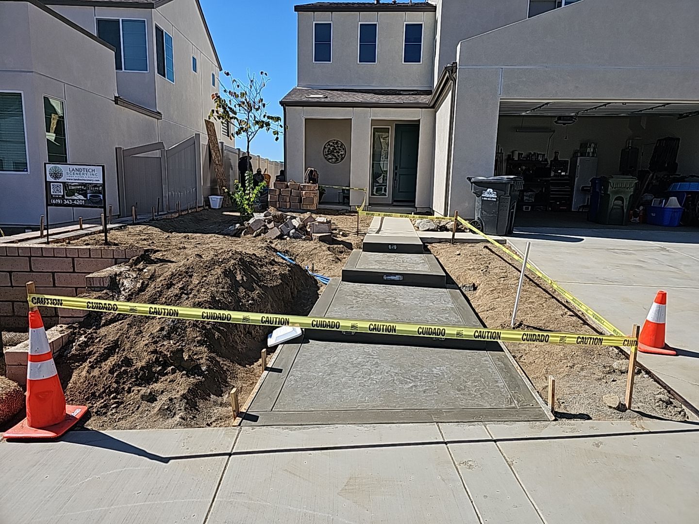 Concrete walkway under construction, house in background. Dirt piles, caution tape, and orange cones present.