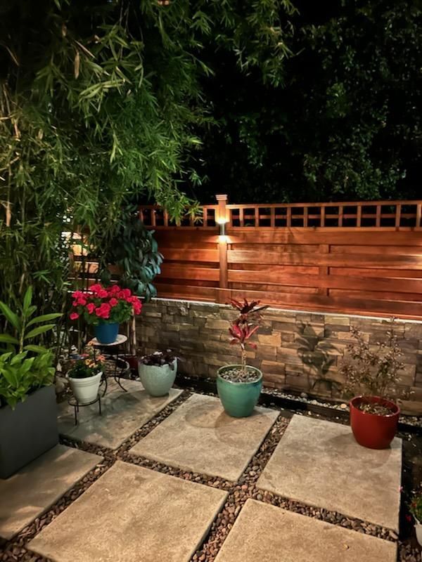 Night view of a patio with stone pavers, potted plants, and a wooden fence, illuminated by ambient lighting.
