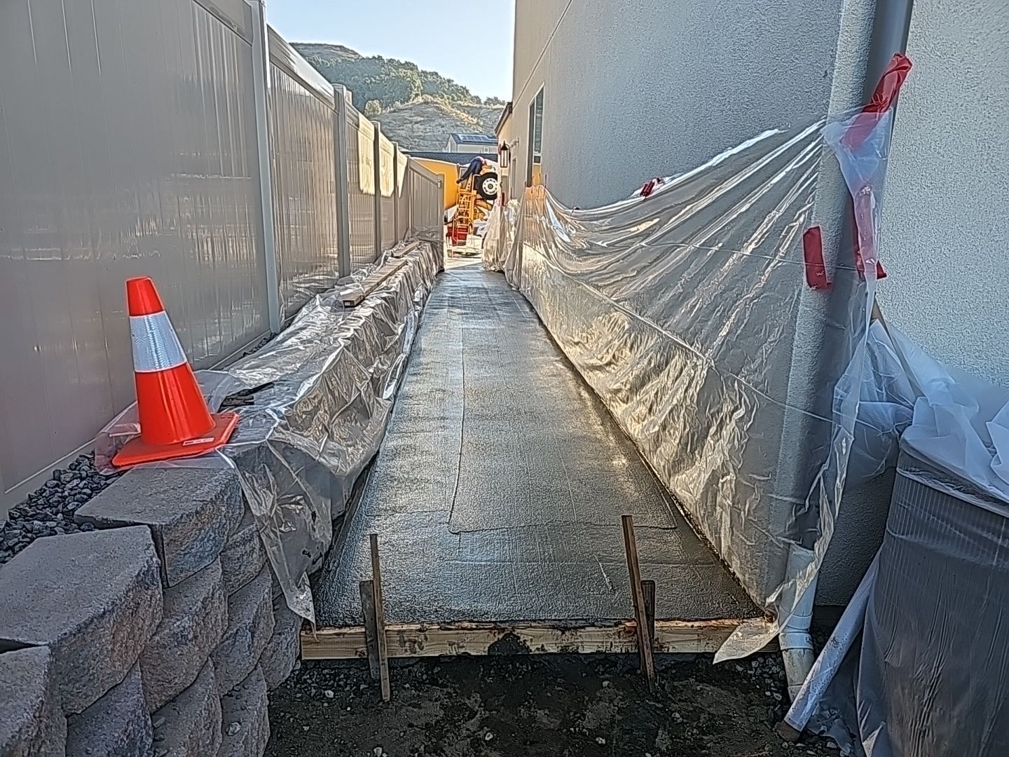 Narrow concrete walkway under construction; orange cone, retaining wall, tarp, and building walls present.