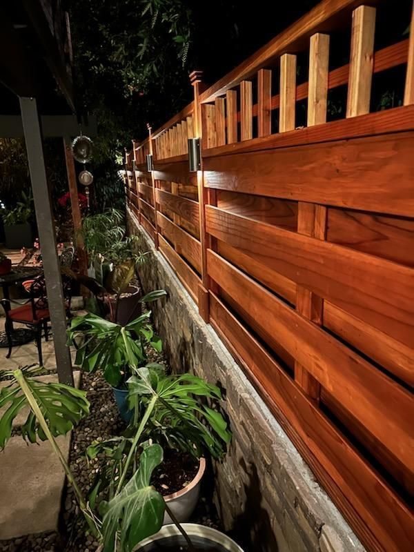 Wooden fence with built-in lighting beside a row of potted plants along a wall.