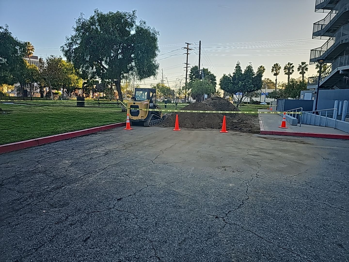 Construction site: Small excavator digs dirt next to a paved area, with orange cones and caution tape.