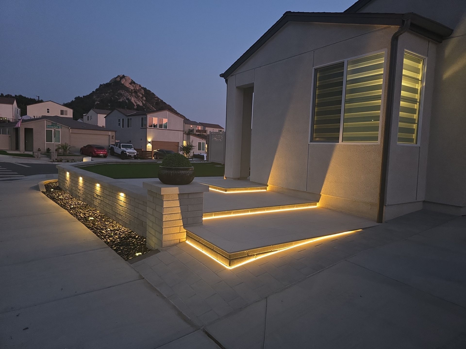Lit exterior of a home at dusk with step and wall lighting, steps leading to the front door, other houses in the background.